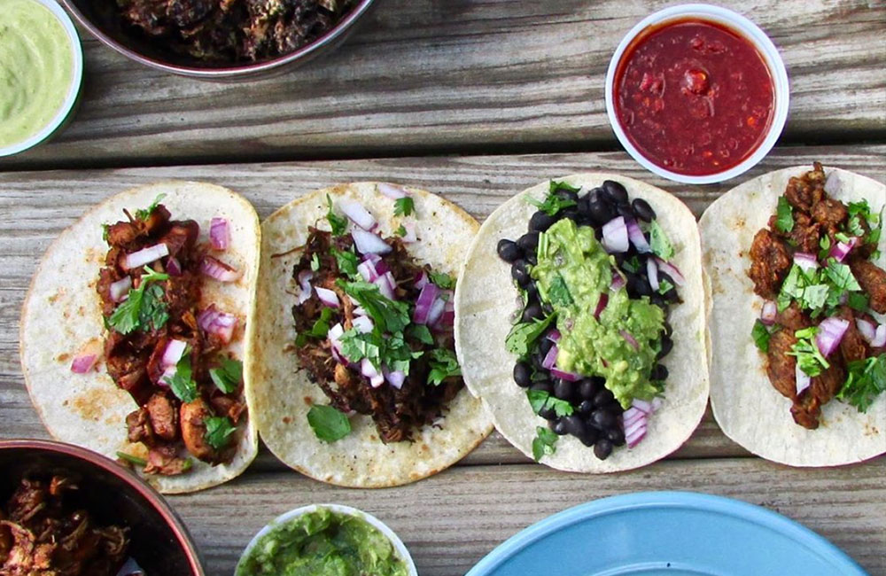 Four soft shell tacos from Dos Amigos sitting on a picnic table with condiments