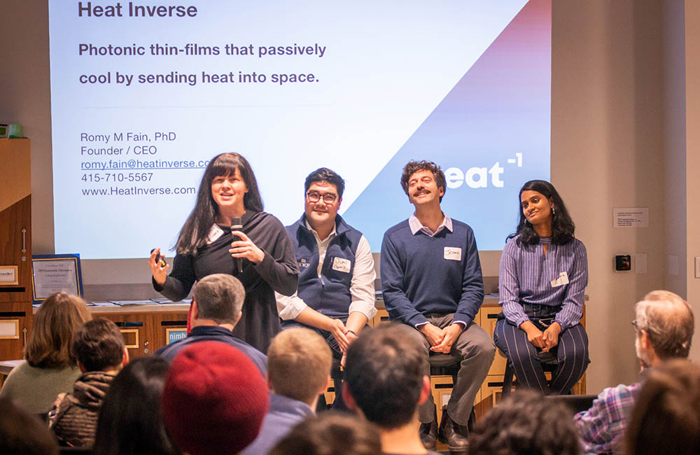CEO and founder of Heat Inverse, Romy Fain, holds a microphone and speaks to an audience in front of a projected presentation on her company while three other panelists watch from the background.