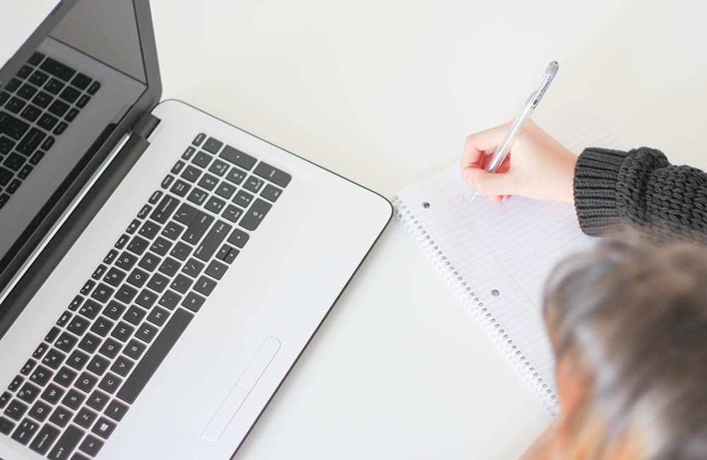 A person takes notes in a notebook while watching a webinar on a laptop.