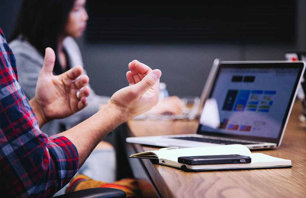 Hands gesturing in front of a laptop in the foreground while a person works on a laptop in the background.