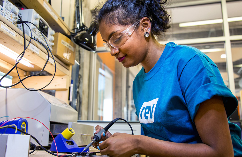 A person wearing safety goggles and a blue Rev tee shirt sodders a piece of hardware.