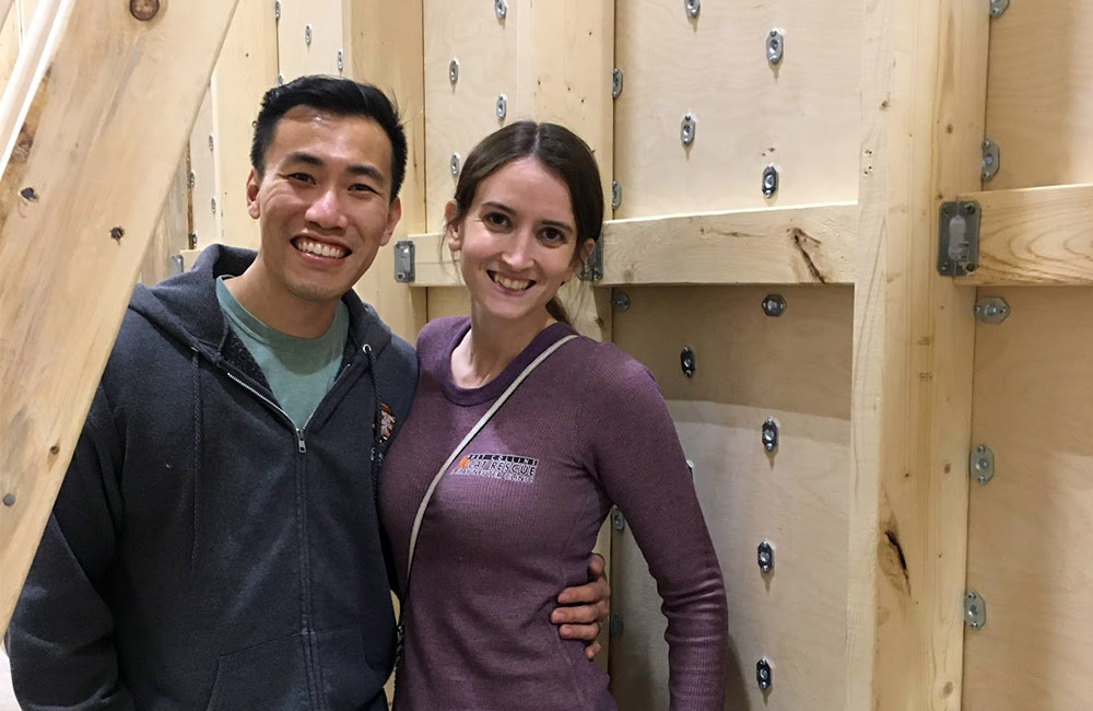 Keith Liao and Beth Rosenliao stand smiling in front of a newly built bouldering wall at their climbing gym. 