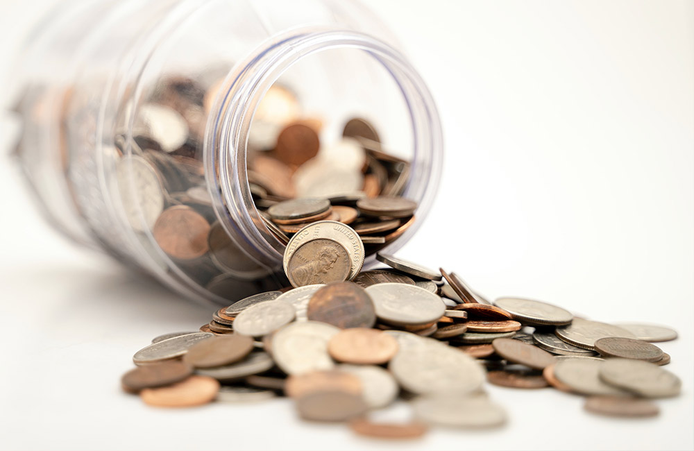 Coins spill out of a jar tipped on its side. The jar and coins are on a white surface and background.