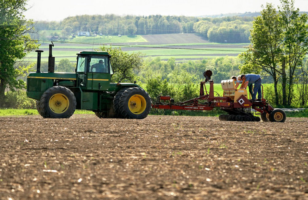A tractor pulls a planter in a freshly sowed field. Three people stand on the planter to load chickpeas into it.