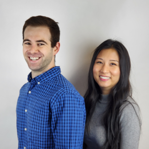 White man and Asian woman stand together in front of a white wall